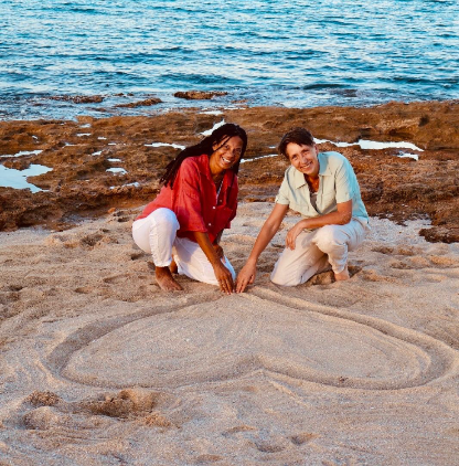 Carmen und Manuela, Gründerinnen von Essence of Love, lachen am Strand und malen ein Herz in den Sand – sie freuen sich darauf, euch mit freien Trauungen, authentischer Fotografie und Lichtbriefen zu begleiten